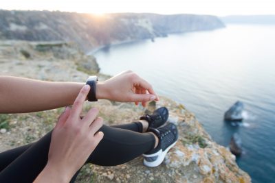 girl on cliff edge by water pointing to fitness tracker on wriist