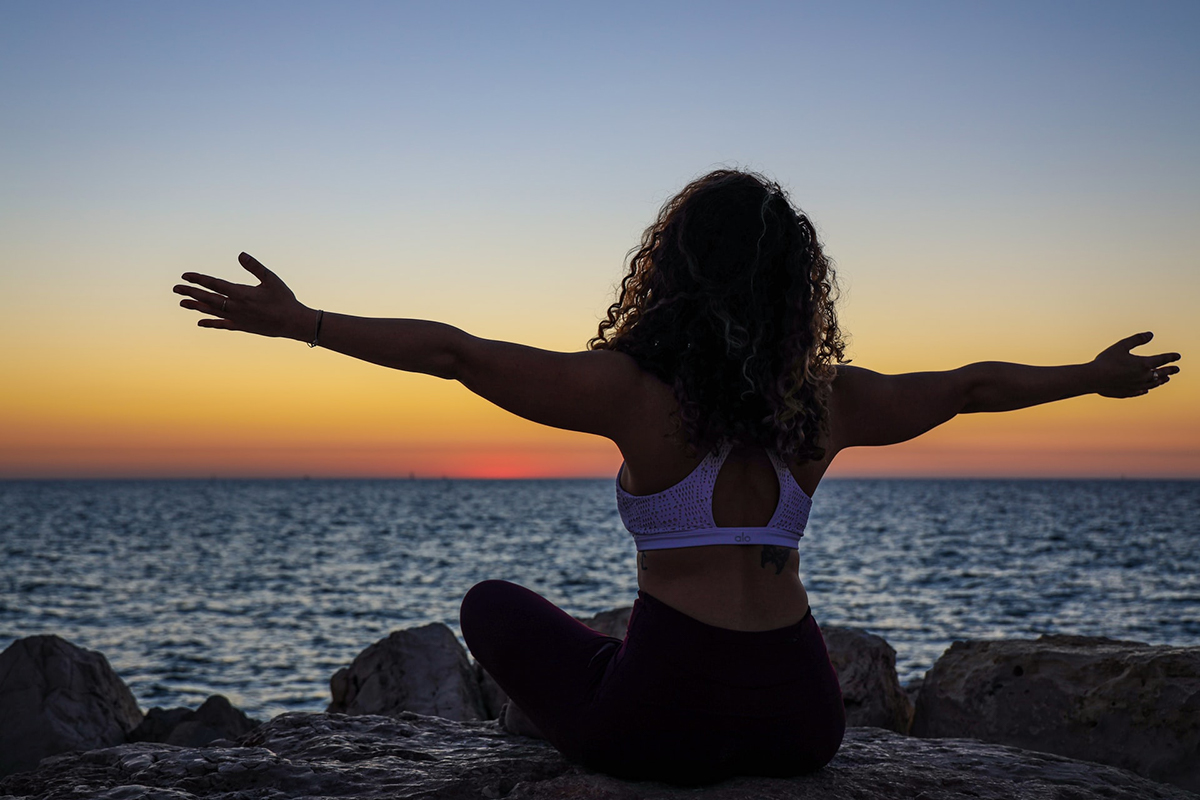 Woman meditating on rocks beside ocean. Photographed by Mor Shani. Image via Unsplash