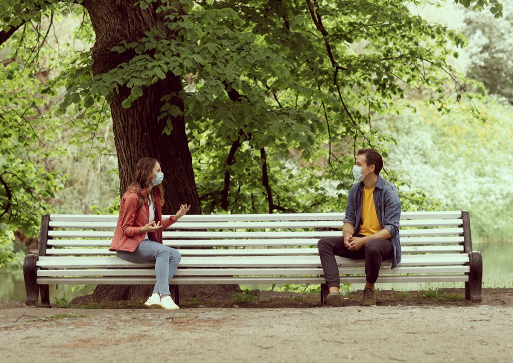 Couple sitting 2 metres apart on park bench to comply with COVID-19 social distancing rules. Image via shutterstock