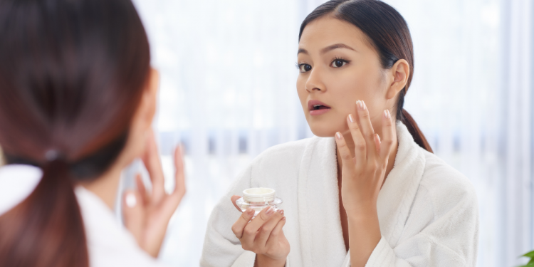 Woman putting on moisturiser in the mirror. Photographed by Dragon Images.