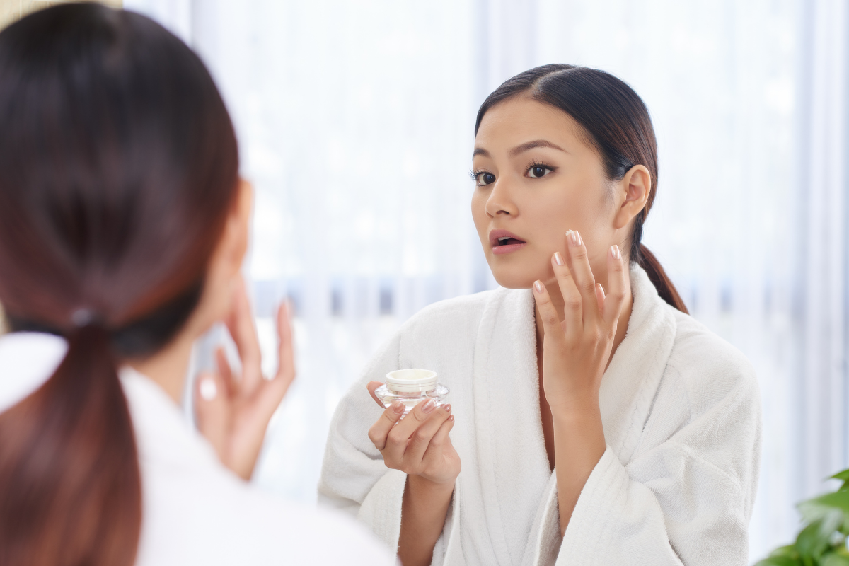 Woman putting on moisturiser in the mirror. Photographed by Dragon Images.