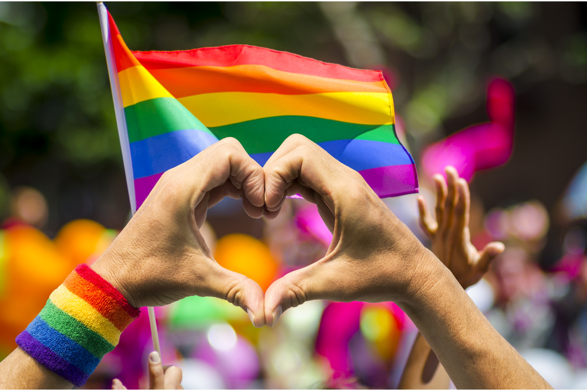 Heart hand gesture against a queer flag. Photography by lazyllama. Image via Shutterstock