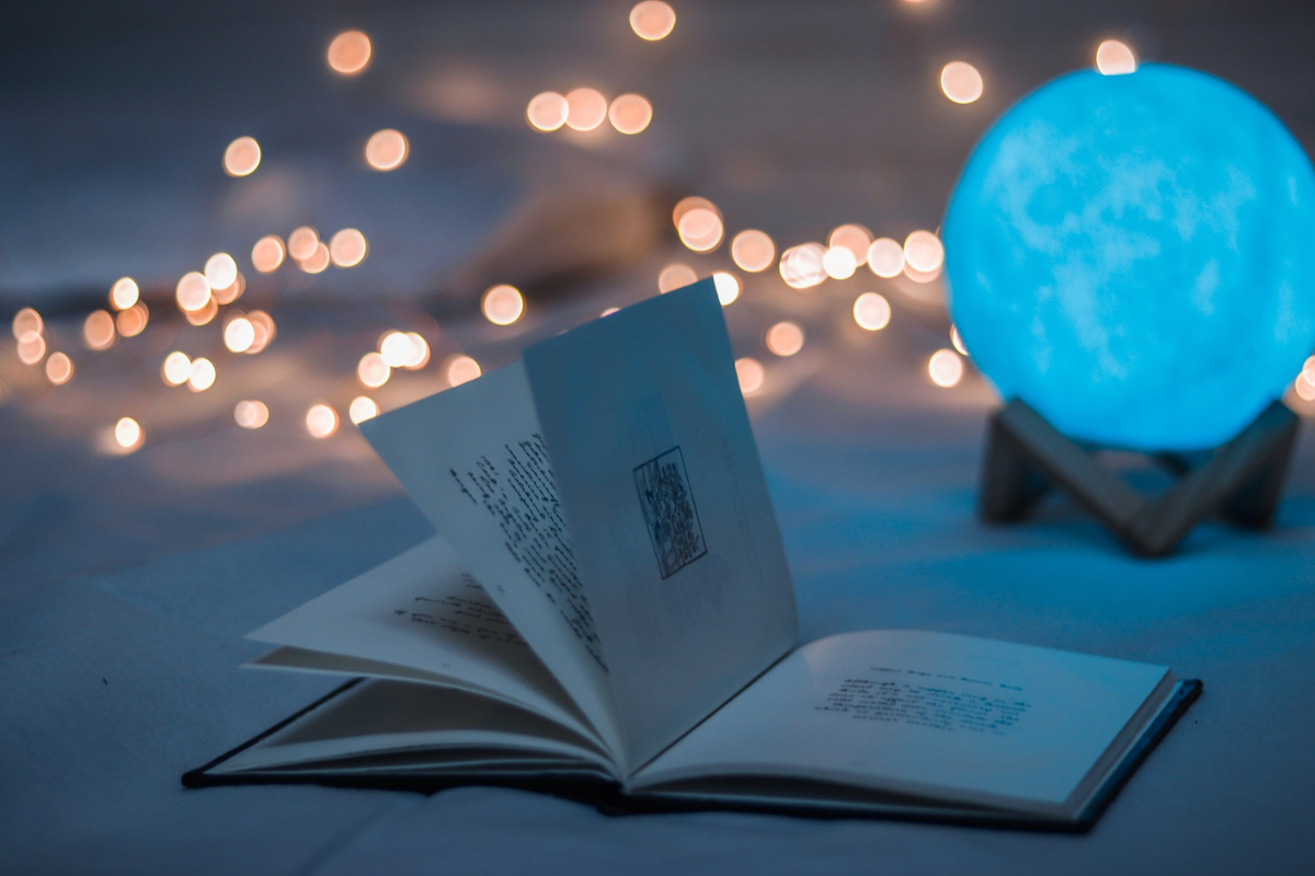 Book open on bed, with fairy lights and orb in background. Photography by Dollar Gill. Image via Unsplash